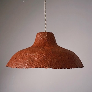 Pode incluir: Uma luminária pendente com um abajur castanho-avermelhado texturizado. O abajur tem uma forma de tigela larga e rasa e é suspenso por um cabo torcido branco. A luminária foi projetada para uso interno.