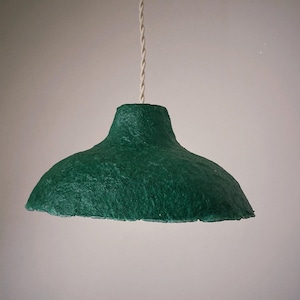May include: A handcrafted, green pendant light fixture with a textured surface. The lampshade has a wide, rounded shape and is suspended by a white, twisted cord. The light fixture is designed for indoor use.