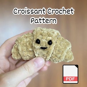 May include: A tan, plush, croissant-shaped crochet item with two small black plastic eyes and a stitched black mouth. The text "Croissant Crochet Pattern" is displayed above the item, and a white PDF icon is visible in the bottom corner.