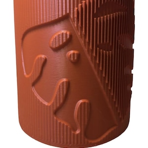May include: A matte rust colored cylindrical container with a textured surface and a raised design of a stylized animal head on the front.