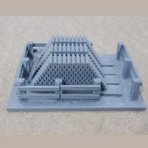 May include: A grey 3D printed model of a wooden crate filled with stacked cannonballs. The crate is on a grey base.