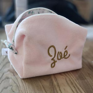 Può includere: Un beauty case rosa chiaro in tessuto felpato con chiusura a zip. Il nome "Zoé" è ricamato in oro. L'interno è bianco con un motivo floreale verde. La borsa ha un manico.
