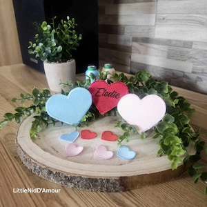 Peut inclure: Une plaque en bois en forme de cœur avec une variété de cœurs en feutre de différentes couleurs, dont le rouge, le rose et le bleu. Le cœur rouge a le nom "Elodie" brodé dessus.