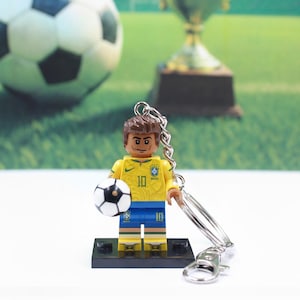 May include: A yellow and blue soccer player figurine keychain holding a soccer ball. The figurine is wearing a yellow jersey with the number 10 and blue shorts. The keychain has a silver metal ring and clasp.