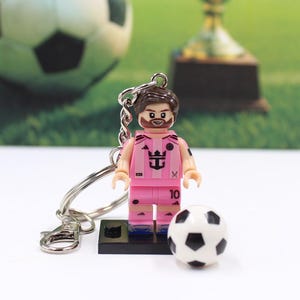 May include: A pink soccer player figurine keychain with a soccer ball. The figurine has a beard and is wearing a pink uniform with the number 10. The keychain is attached to a silver metal ring.