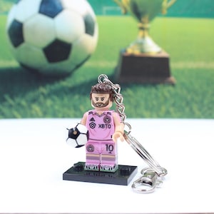 May include: A pink and white soccer player figurine keychain holding a soccer ball. The figurine has the number 10 on the jersey and the text "XBTO". A soccer ball and trophy are in the background.