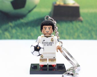 May include: A Lego-style keychain featuring a soccer player in a white uniform with the text "Emirates FLY BETTER." The player holds a black and white soccer ball. The keychain has a silver metal ring and clasp.
