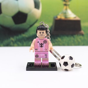 May include: A pink soccer player figurine keychain with a small soccer ball charm. The figurine is wearing a pink uniform with the number 9. The keychain is on a black base, with a soccer ball and trophy in the background.