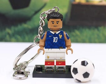 May include: A keychain featuring a plastic minifigure of a soccer player wearing a blue jersey with the number 10, white shorts, and red accents. The figure stands on a black base next to a small white and black soccer ball, attached to a metal ring.