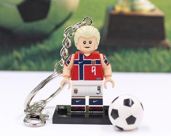 May include: A keychain featuring a blonde minifigure wearing a red and blue Norway soccer jersey with the number 9, white shorts with the number 9, and a small black base. A miniature black and white soccer ball sits next to the minifigure.