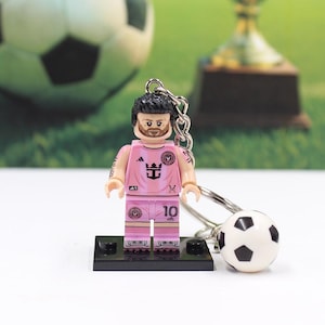 May include: A pink soccer player figurine keychain with the number 10 on the uniform. The keychain includes a small soccer ball charm. The figurine is on a black base. A soccer ball and trophy are in the background.