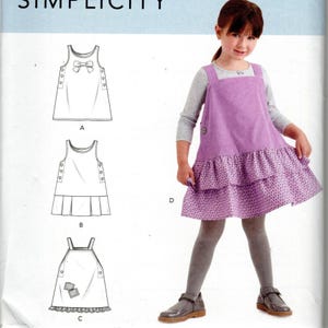 Puede incluir: Un sobre de patrón de costura Simplicity con ilustraciones de tres variaciones de vestido etiquetadas como A, B y C. La imagen muestra a un niño con un vestido tipo pichi morado con falda de dos volantes y una camiseta gris de manga larga.