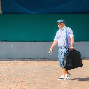 May include: A man wearing a light blue collared shirt, blue jeans, and a blue cap walks down a brick walkway while carrying a black garment bag.