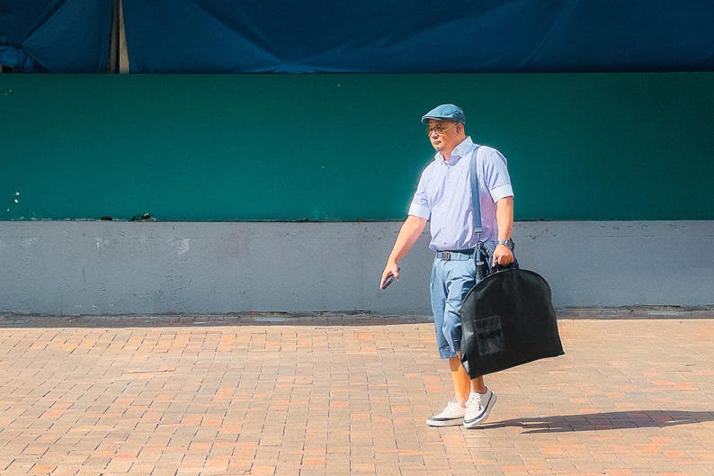 May include: A man wearing a light blue collared shirt, blue jeans, and a blue cap walks down a brick walkway while carrying a black garment bag.