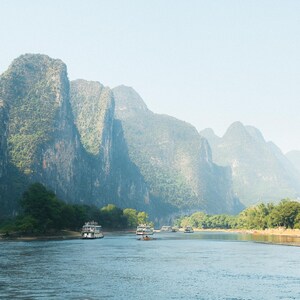 May include: A scenic view of a river with several boats traveling through a mountainous landscape. The mountains are covered in lush green foliage and the water is calm and still.