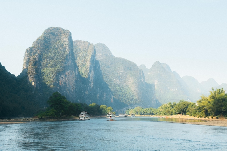 May include: A scenic view of a river with several boats traveling through a mountainous landscape. The mountains are covered in lush green foliage and the water is calm and still.
