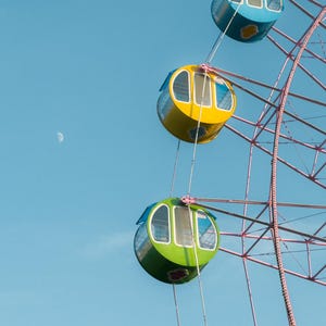May include: A pink Ferris wheel with colorful gondolas against a blue sky with a crescent moon. The gondolas are yellow, green, and blue.
