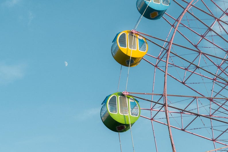 May include: A pink Ferris wheel with colorful gondolas against a blue sky with a crescent moon. The gondolas are yellow, green, and blue.