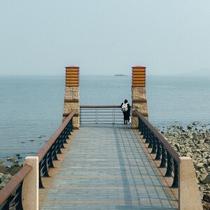 May include: A person stands on a wooden walkway overlooking a body of water. The walkway is lined with black railings and leads to two stone pillars with orange tops.