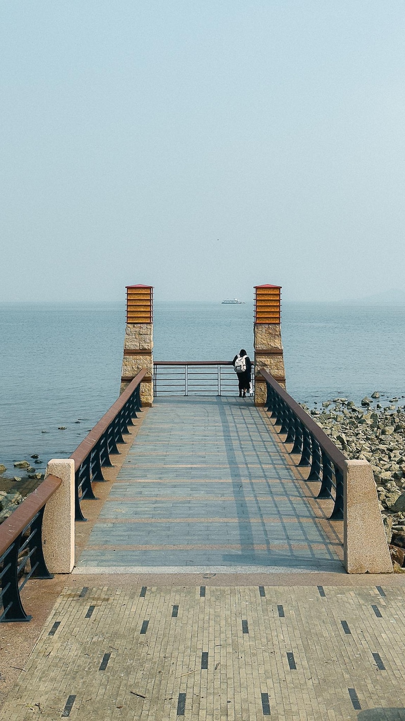May include: A person stands on a wooden walkway overlooking a body of water. The walkway is lined with black railings and leads to two stone pillars with orange tops.