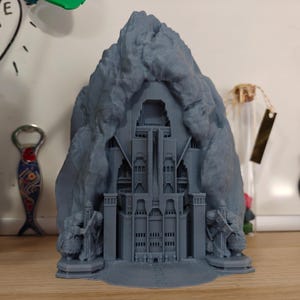 May include: A gray 3D-printed model of a fortress nestled within a mountain. The detailed structure features towers, archways, and figures at the base. The model is displayed on a wooden surface.