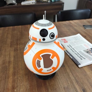 Op de afbeelding: Een wit en oranje Star Wars BB-8 droid speelgoed met een zwart oog en een grijze bovenkant. De droid staat op een houten tafel.