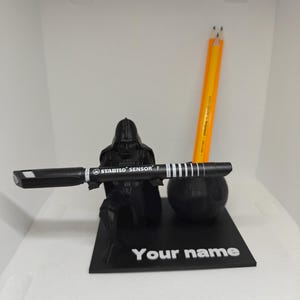 Könnte beinhalten: Ein schwarzer Schreibtisch-Organizer mit einer Darth-Vader-Figur, die einen schwarzen Stift mit dem Text "STABILO SENSOR F" hält. Enthält ein Todesstern-Modell und zwei gelbe Bleistifte. Die Basis lautet "Your name".