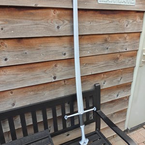May include: A long, silver sword with a cross-shaped hilt and a black handle rests on a black bench. The sword is displayed against a backdrop of wooden planks and a sign that reads "MAN CAVE".