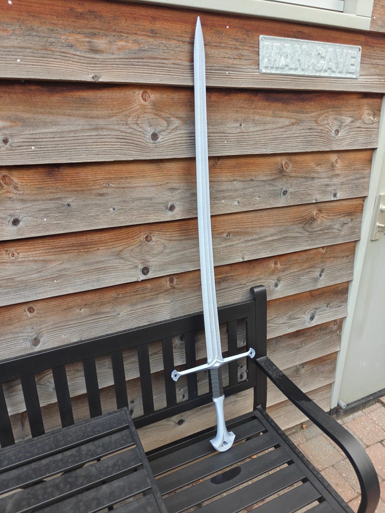 May include: A long, silver sword with a cross-shaped hilt and a black handle rests on a black bench. The sword is displayed against a backdrop of wooden planks and a sign that reads "MAN CAVE".