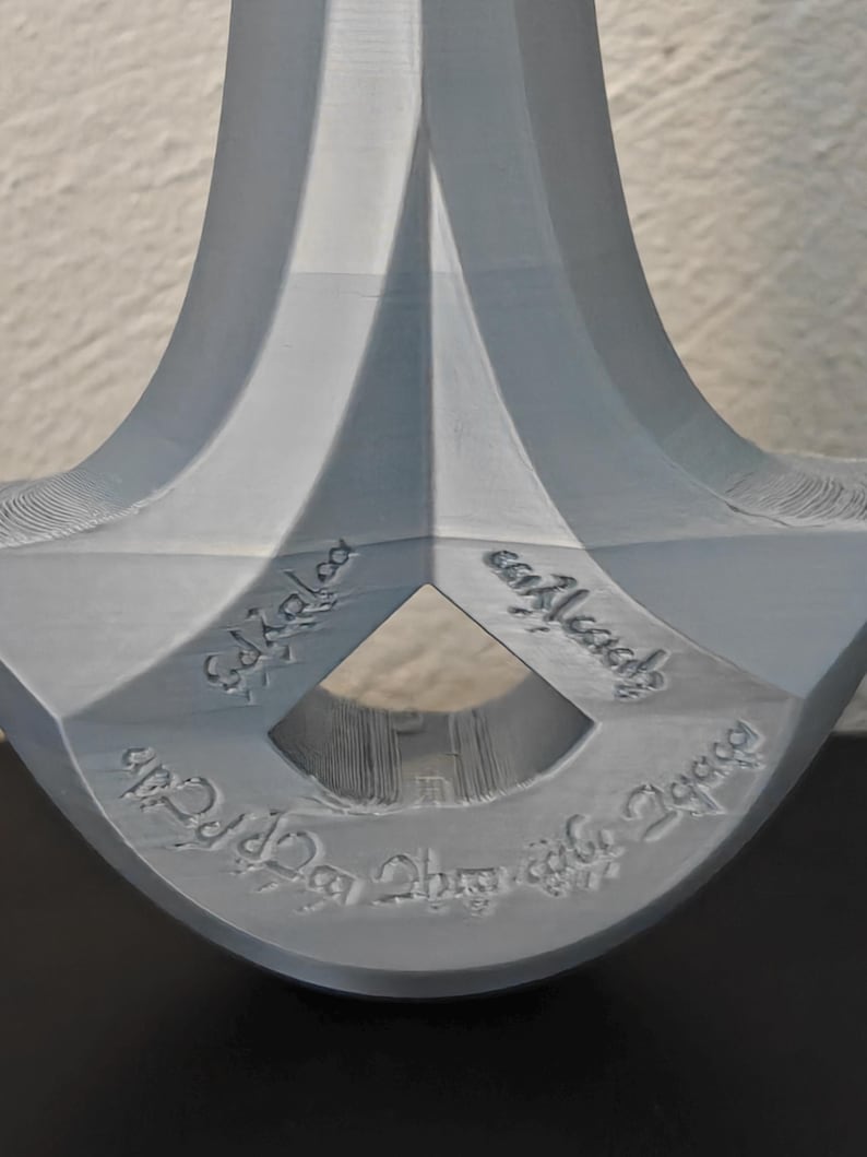 May include: A close-up of a gray, stylized sword hilt with engraved text. The hilt has a diamond-shaped opening and a curved design. The text is in a stylized font.