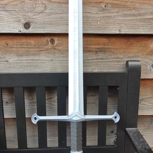 May include: A silver-colored sword with a long, engraved blade and a crossguard. The hilt is gray with a silver pommel. The sword is resting on a black bench with a wooden background. The blade has text engraved on it.