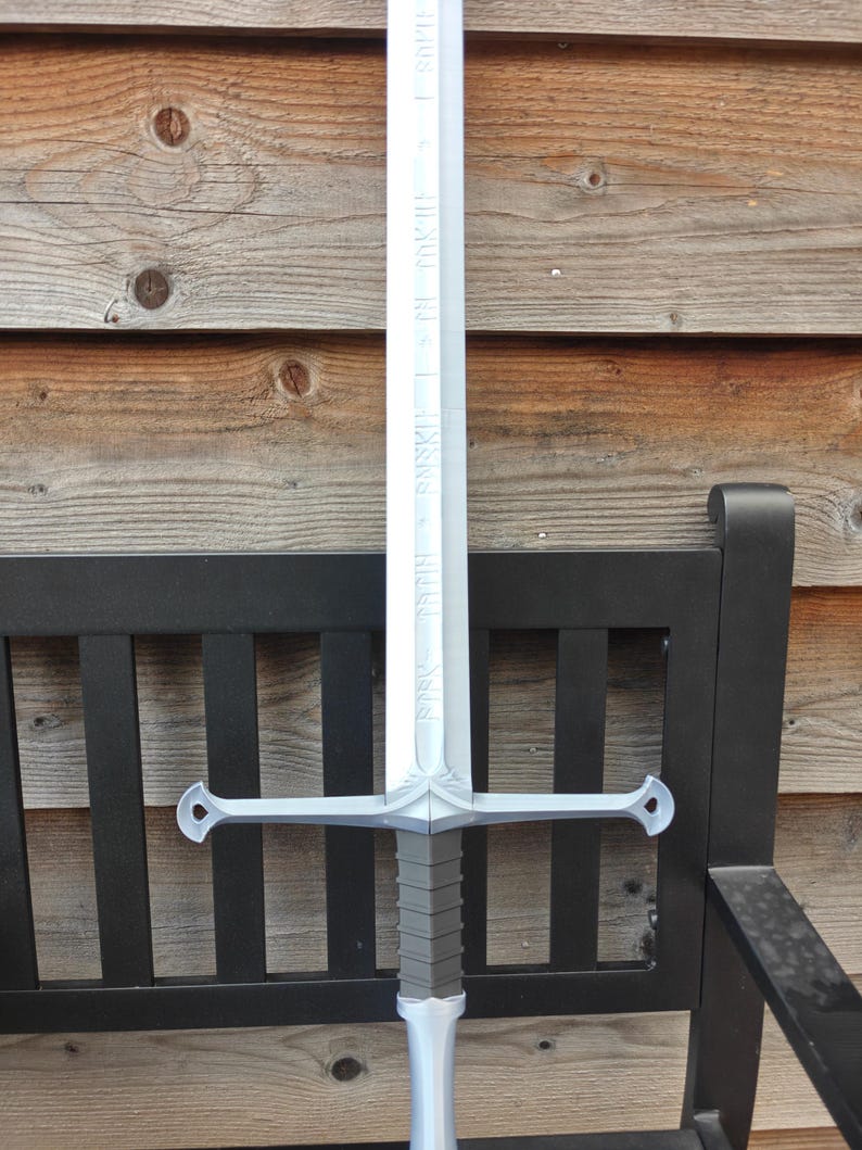 May include: A silver-colored sword with a long, engraved blade and a crossguard. The hilt is gray with a silver pommel. The sword is resting on a black bench with a wooden background. The blade has text engraved on it.