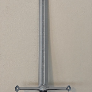 May include: A silver-colored sword with a black handle. The blade has etched inscriptions and a crossguard with rounded ends. The sword is displayed against a neutral-colored wall.