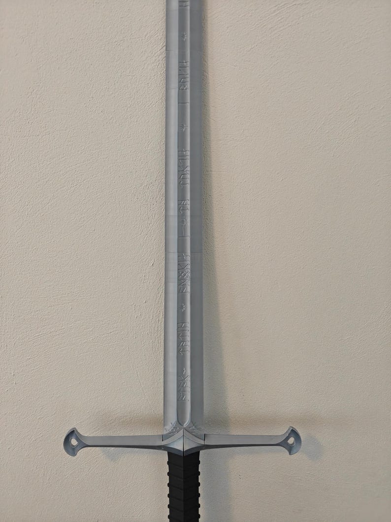 May include: A silver-colored sword with a black handle. The blade has etched inscriptions and a crossguard with rounded ends. The sword is displayed against a neutral-colored wall.