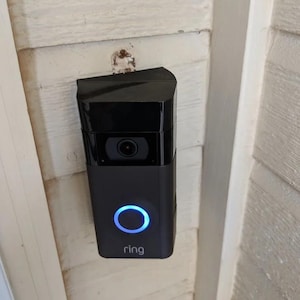 Puede incluir: Un timbre Ring negro con un anillo de luz azul alrededor del botón. El timbre está montado en una pared blanca.