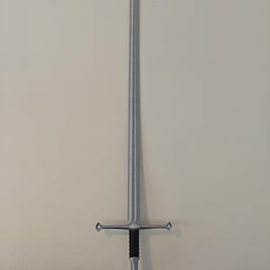 May include: A silver-colored sword with a long, pointed blade and a cross-shaped hilt. The handle is black with a silver pommel. The sword is displayed against a neutral-colored wall.