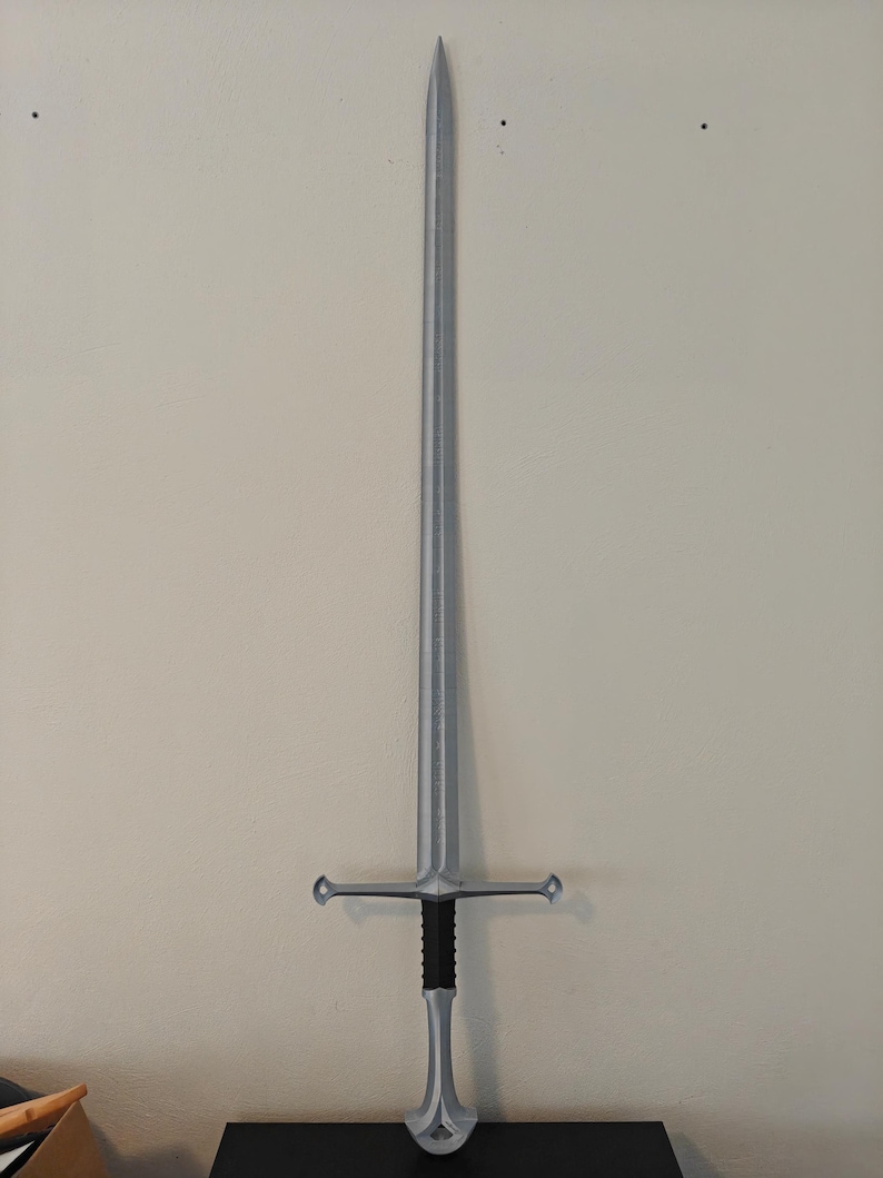 May include: A silver-colored sword with a long, pointed blade and a cross-shaped hilt. The handle is black with a silver pommel. The sword is displayed against a neutral-colored wall.