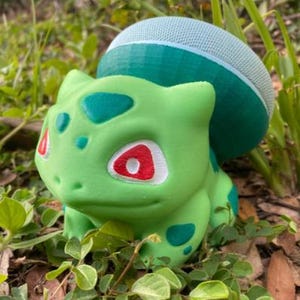 May include: A green and blue 3D printed figurine of a Pokemon character, Bulbasaur. The figurine has a light blue speaker on its head.