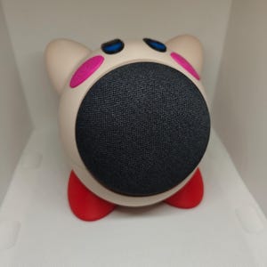 Peut inclure: Un haut-parleur intelligent en forme d'animal blanc et rose avec une grille de haut-parleur en tissu noir et des pieds rouges.