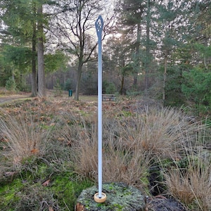 Op de afbeelding: Een lange, slanke, witte verticale toverstaf met een ingewikkeld, opengewerkt filigraanontwerp aan de bovenkant. De basis van de staf zit in een kleine, ronde, lichtgekleurde houten standaard die op een bemoste boomstronk rust.