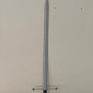 May include: A silver-colored sword with a long, straight blade and a cross-shaped hilt. The hilt has a black grip and a decorative design. The sword is displayed against a neutral-colored wall.