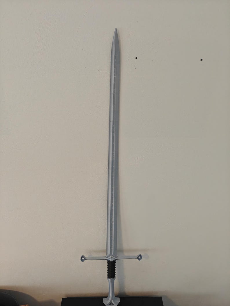 May include: A silver-colored sword with a long, straight blade and a cross-shaped hilt. The hilt has a black grip and a decorative design. The sword is displayed against a neutral-colored wall.