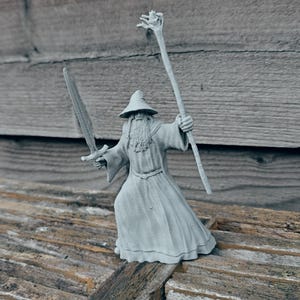 May include: A gray 3D printed figurine of a wizard holding a staff and a sword. The wizard is wearing a long robe and a pointed hat.
