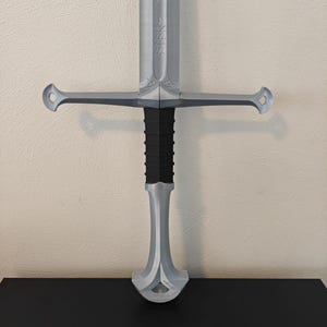 May include: A silver-colored sword with a black handle. The sword features a crossguard with rounded ends and engraved details on the blade. The handle has a textured, ribbed design.