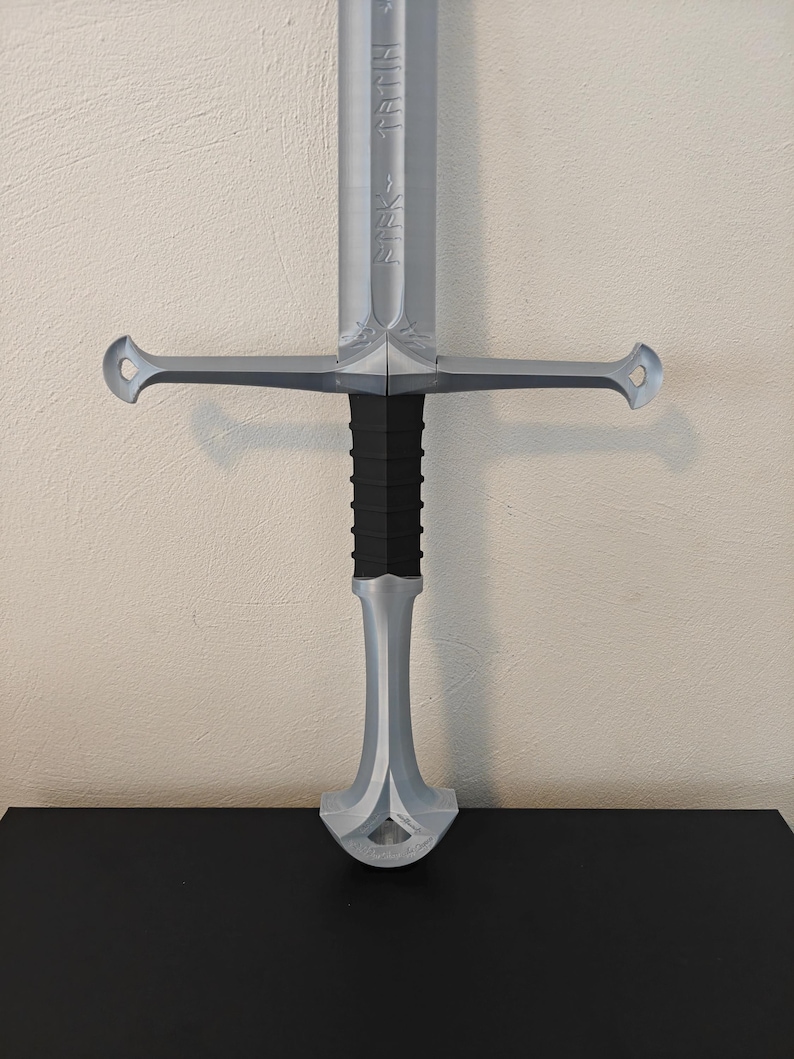 May include: A silver-colored sword with a black handle. The sword features a crossguard with rounded ends and engraved details on the blade. The handle has a textured, ribbed design.