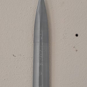 May include: A gray, bullet-shaped object with a pointed tip. The object has vertical grooves and the text "BAKEAKHA" and "BAH" printed on its side. The object is set against a neutral-colored wall.