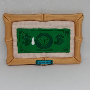 May include: A decorative piece featuring a green dollar bill design with a seashell and tear drop graphic. The dollar bill is set within a gold-colored frame, with the text "FIRST DOLLAR" on a blue bar at the bottom.