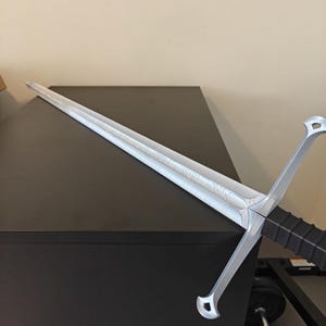 May include: A silver-colored sword with a long, straight blade and a crossguard. The hilt is black with a textured grip. The blade has etched detailing. The sword is displayed on a dark surface.