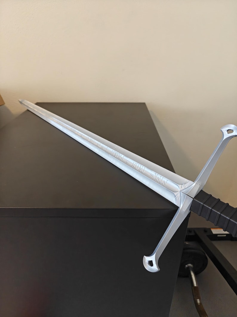 May include: A silver-colored sword with a long, straight blade and a crossguard. The hilt is black with a textured grip. The blade has etched detailing. The sword is displayed on a dark surface.