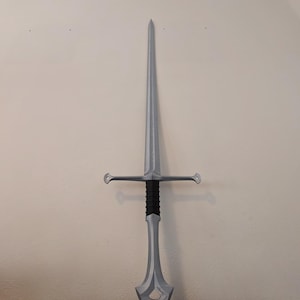 May include: A silver-colored sword with a long, pointed blade and a crossguard. The handle is black and gray, and the sword is displayed against a white wall. The sword's overall design suggests a fantasy or historical theme.